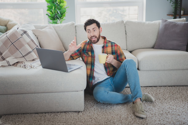 漂亮的年轻人的全尺寸照片坐在地板上视频通话上网本喝茶穿时髦的格子衬衫现代光室内平室内图片下载