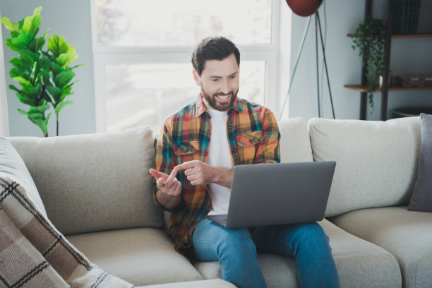英俊的年轻人的照片坐在沙发上网本视频电话穿着格子衬衫度过休闲宽敞的房子在室内房间图片下载