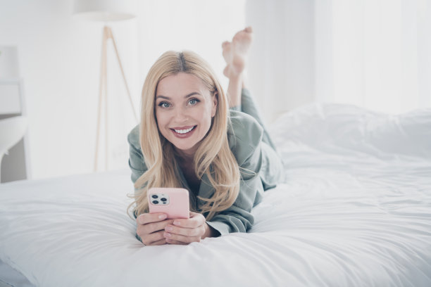 漂亮的年轻女子躺在床上的全尺寸照片使用电话睡衣白色室内卧室公寓室内图片下载