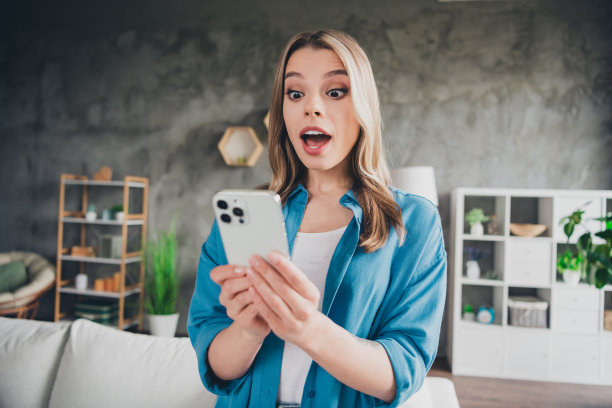 照片中，年轻的商务女士穿着蓝色衬衫，惊讶地看着智能手机，看着她的阁楼办公室，为客户提供不断增长的房地产价格图片下载