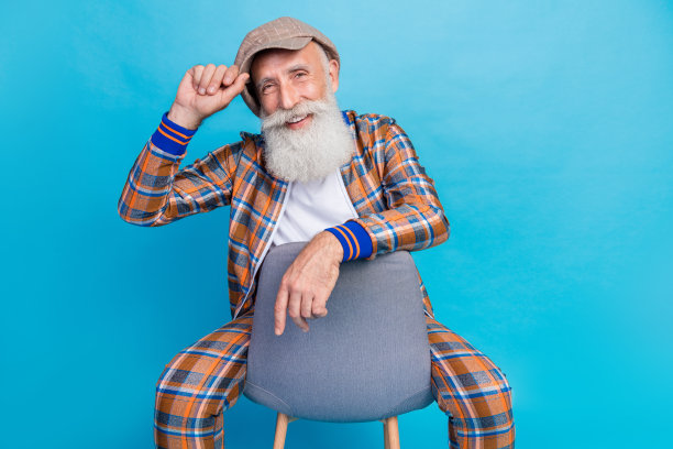 照片自信英俊的男子养老金领取者穿着格子服装坐在椅子手臂头饰孤立的蓝色背景图片下载