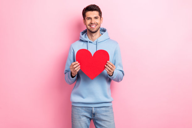 照片中开朗可爱的小伙子穿着蓝色运动衫，拿着红色的心微笑着孤立的粉红色背景图片下载