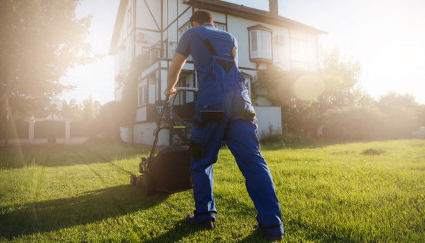 在一个阳光明媚的日子里，一名身穿蓝色工作服的男子正在一所房子前修剪草坪，灯光照亮了现场。这张照片捕捉到了一个宁静的、保存完好的花园，散发着温暖的金色光芒图片下载