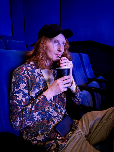 特写照片中，红头发的男人留着长长的波浪发型，穿着休闲服，有图案的夹克，黑色牛仔牛仔裤，戴着深蓝色的帽子，拿着一次性饮料杯，用吸管喝水，这是前景的焦点图片下载