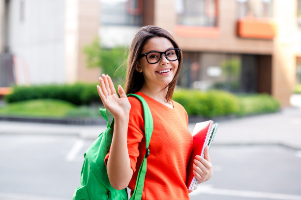 照片漂亮的青少年好心情的女孩抄写书挥手hi学生穿红色的衣服走在城市公园外图片下载