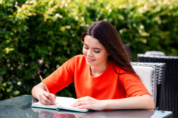 照片漂亮的青少年好心情的女孩坐在桌子咖啡馆写笔记本穿着红色的衣服走在城市城市公园外图片下载