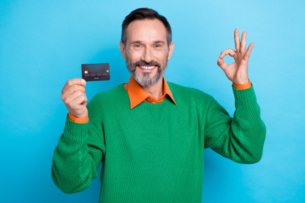 照片积极的好心情，微笑的专业银行家穿着绿色的套头衫，显示ok的标志，拿着塑料卡促销孤立在蓝色的背景图片下载