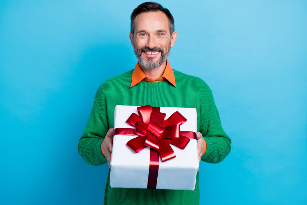 成熟的英俊男子手持大礼盒庆祝红色蝴蝶结，穿着时髦的绿色服装，与蓝色背景隔离图片下载