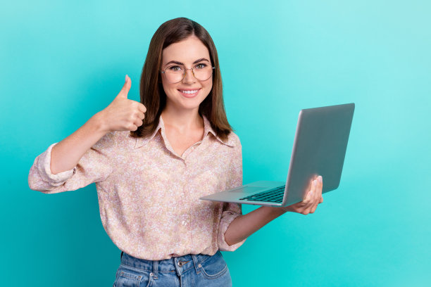 照片中，甜美自信的女孩穿着粉色衬衫，戴着眼镜，拇指朝上聊天，现代设备孤立的蓝绿色背景图片下载