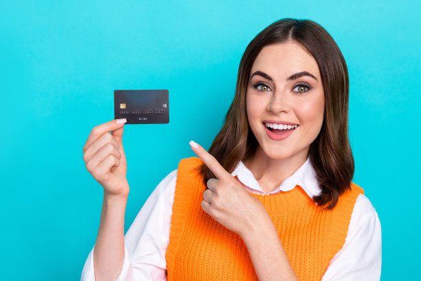 照片中开朗满意的可爱女子棕色头发，穿着橙色马甲，对着蓝绿色背景上孤立的塑料卡片图片下载