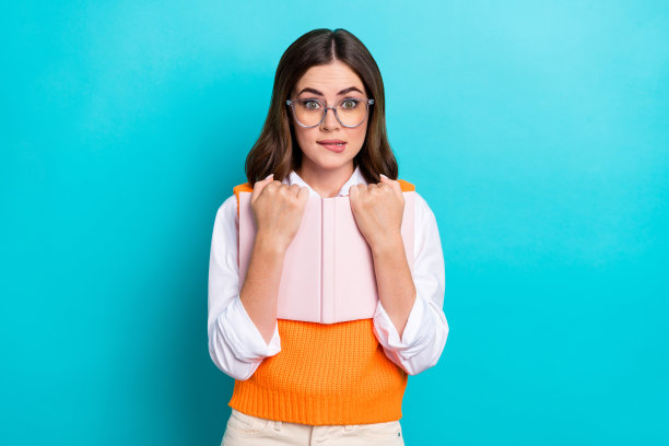 照片正面开朗印象深刻的女人直发穿橙色马甲拥抱书咬嘴唇孤立在蓝绿色的背景图片下载