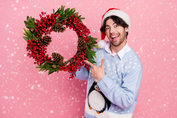 照片上的年轻人穿着蓝色的圣诞套衫，手指着花环，微笑着孤立的粉红色背景图片下载