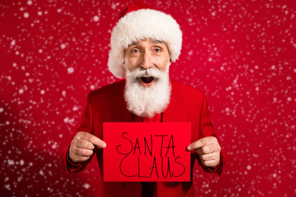 照片中迷人的成熟男子穿着圣诞服装，帽子拿着圣诞老人标牌孤立的红色背景图片下载