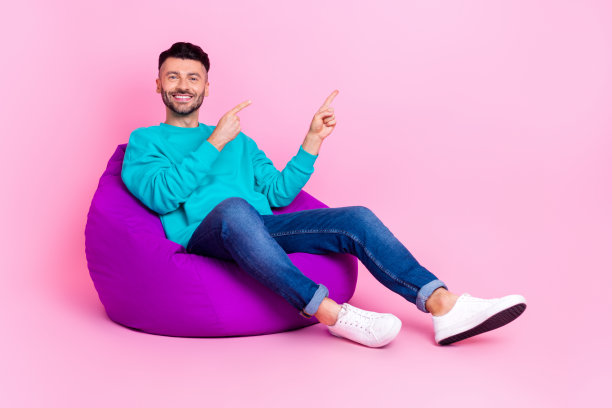 全尺寸照片的年轻帅哥坐在柔软的豆袋点空空间穿着时尚的蓝色衣服孤立在粉红色的背景图片下载