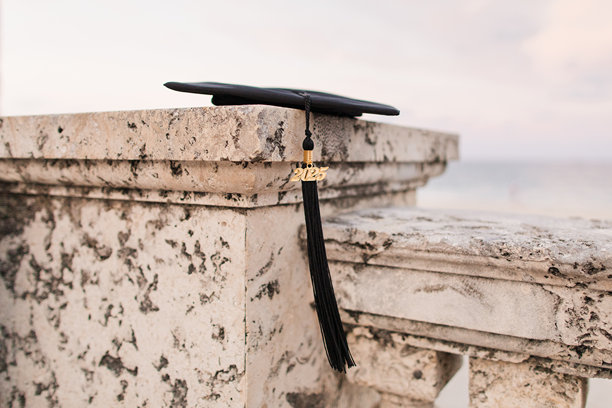 黑色毕业帽和流苏与黄金2025符咒躺在石墙上俯瞰棕榈滩，日落的佛罗里达海岸图片下载