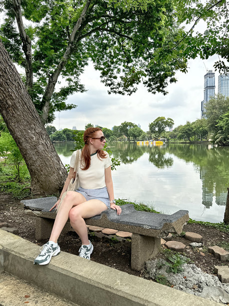 照片中，迷人的红发女子扎着辫子，穿着休闲的短裤、t恤和太阳镜，坐在泰国曼谷帕图姆湾区隆皮尼公园湖滨的混凝土长凳上，背景是城市景观摩天大楼的天际线图片下载