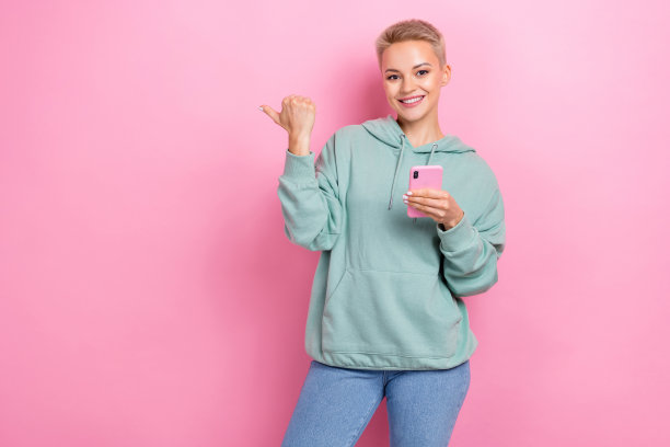 年轻快乐的好心情女孩的照片肖像拿着她的三星手机直接拇指模型新的应用程序购物孤立的粉红色背景图片下载
