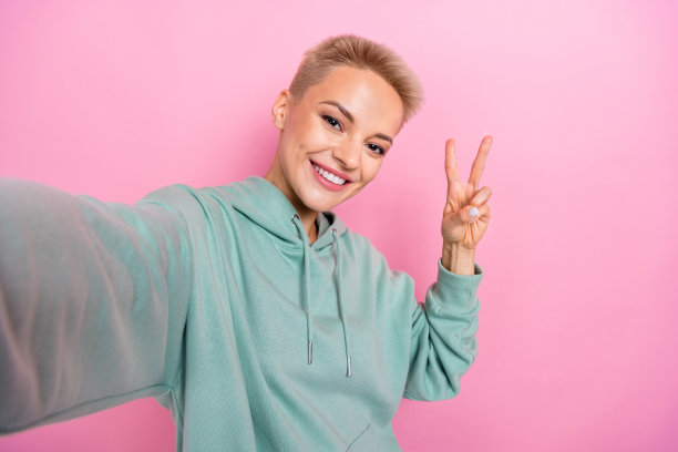 年轻积极满意的金发短发女孩的自拍照片，在粉红色的背景下，在线流媒体上向订阅者展示了v字问候图片下载