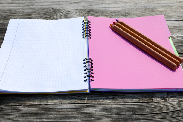 木桌上放着一本练习本，上面有粉红色和白色的书页，还有三支彩色铅笔。垂直的照片。系列回归校园。图片下载