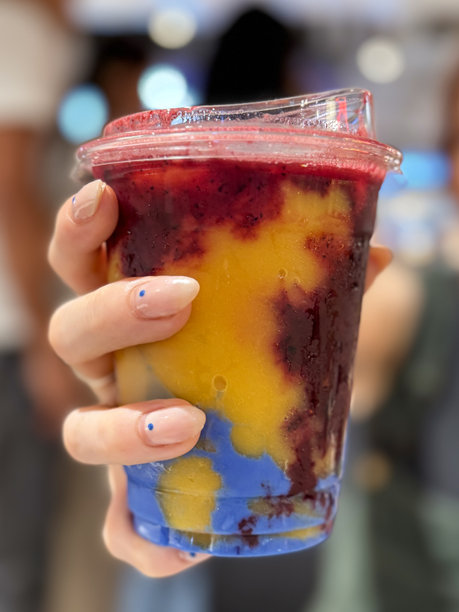 难以辨认的特写图像，穿着绿色灯芯绒工装裤和黑色露脐上衣，拿着塑料的一次性杯盖芒果覆盆子和蓝色螺旋藻奶昔，被照亮的购物中心背景，重点放在前景上图片下载