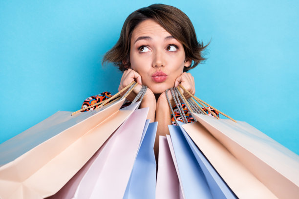 照片中，俏皮可爱的女士穿着印花衬衫，抱着购物者噘起嘴唇，看起来空无一物的蓝色背景图片下载