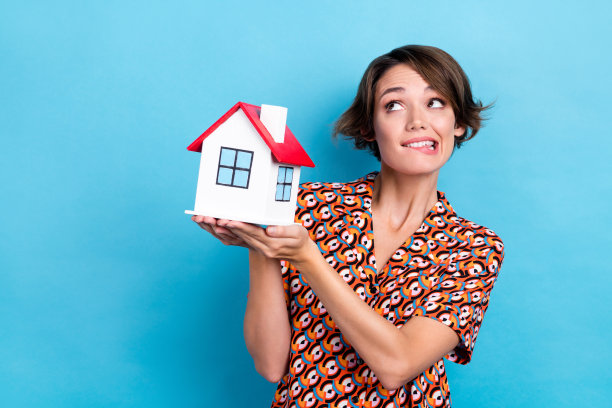 照片的怀疑，不确定的女士穿着印花衬衫咬嘴唇，抱着房子看空空间孤立的蓝色背景图片下载