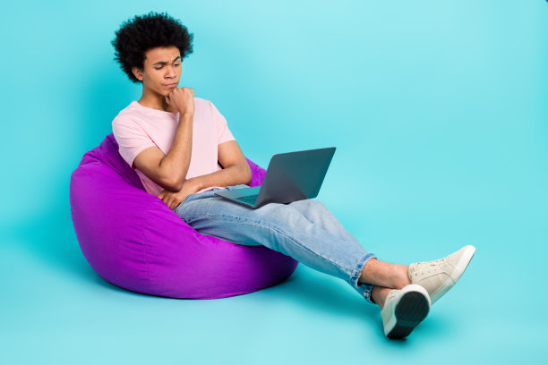 聪明聪明的男人学生的全身照片，沉思着，他的上网本，坐在豆袋里，在青色的背景上共同工作图片下载
