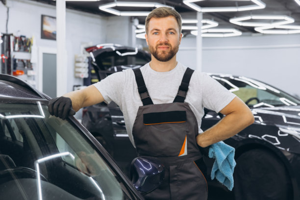 汽车细节，洗车和清洁的概念。留着胡子的年轻男性专业洗车工人戴着黑色橡胶手套，拿着超细纤维擦拭豪车的车身。图片下载