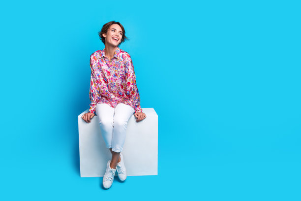 漂亮开朗的女士的全身照片，坐在讲台上的立方体，在蓝色的背景上看起来空荡荡的图片下载