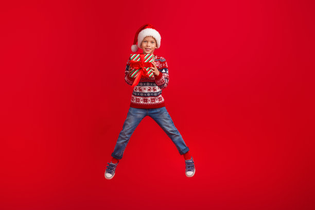 迷人的小男孩跳着拿着礼物庆祝圣诞节穿着针织毛衣孤立的红色背景的全身照片图片下载