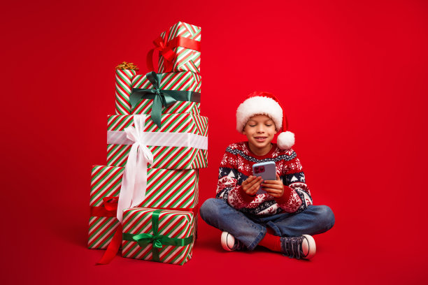 可爱的小男孩的全尺寸照片现在堆抱装置坐在地板上庆祝新年穿着针织服装孤立的红色背景图片下载