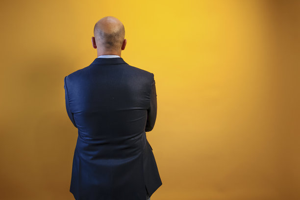 照片中，一名身穿深蓝色西装和浅蓝色衬衫的男子背对着镜头站在一面黄色的墙前。图片下载