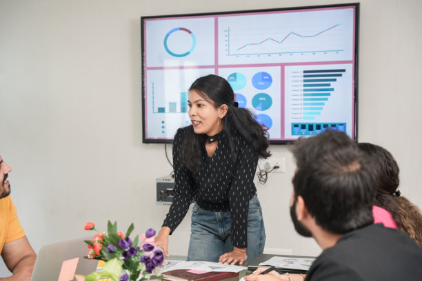 自信的年轻印度妇女在会议室向她的同事做报告或演示。显示图形，饼图和公司的增长在墙上的电视。图片下载