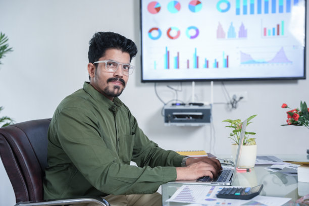 年轻的印度商人或企业家穿着正装坐在办公室里看着镜头的特写。表格上的报告、文件和计算器。创业和财务规划概念。图片下载