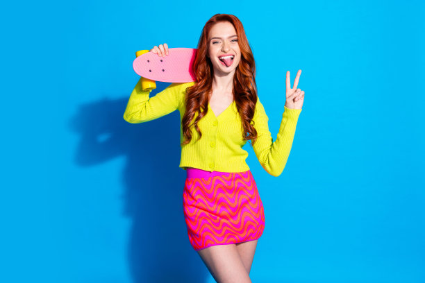 照片肖像迷人的年轻女子舌头出滑板v字穿着时尚的黄色衣服孤立的蓝色背景图片下载