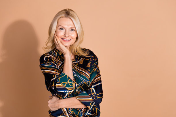 照片肖像的金发成熟年龄的商业女性触摸脸颊在时尚的魅力衬衫孤立的米色背景图片下载