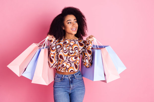 照片中可爱可爱的小女孩穿着印花衬衫抱着购物者望着空旷孤立的粉色背景图片下载