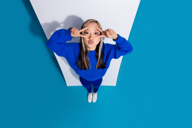 俯视图照片，时髦的韩国女孩靠近手机显示盖脸v标志时髦的符号噘起的嘴唇孤立的蓝色背景图片下载