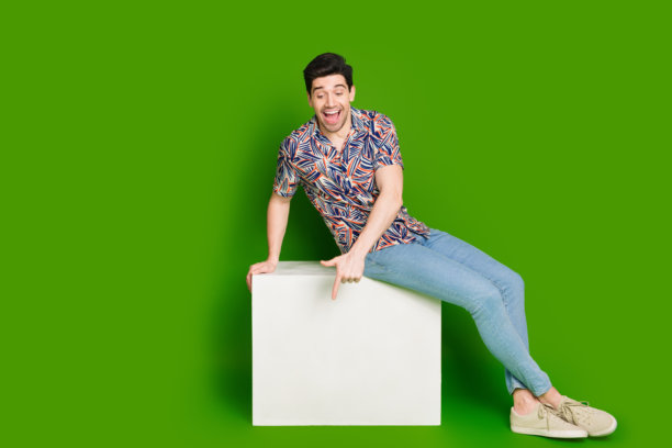 大喜的家伙穿着印花衬衫牛仔裤的全尺寸照片，看着白色的立方体空间孤立在绿色的背景图片下载