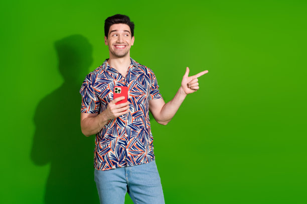 照片中的聪明的家伙，有时尚的发型，穿着印花衬衫，手持智能手机，在绿色的背景上看空的空间图片下载