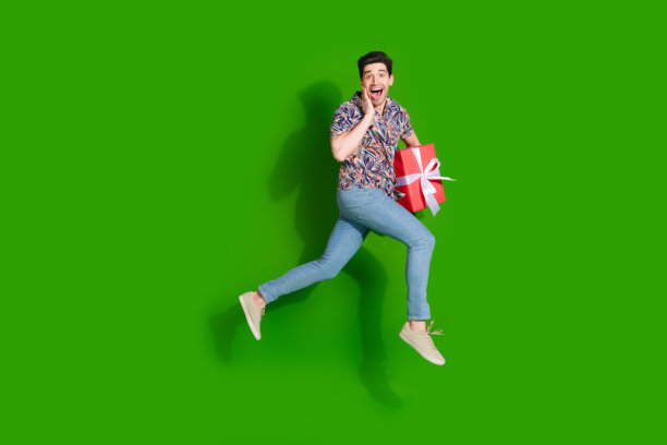 大喜的家伙穿着印花衬衫牛仔裤的全尺寸照片跑到空白的空间，手臂上的礼物盒孤立在绿色背景上图片下载