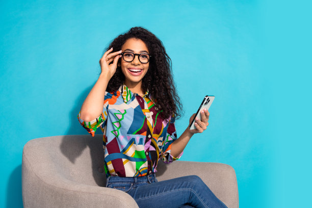 照片正面漂亮可爱的女人穿着时尚的衣服，坐在扶手椅上，戴着眼镜，小玩意孤立的蓝色背景图片下载