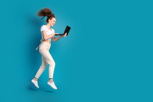 迷人的年轻女子的全身照片拿着上网本跑步跳跃穿着时尚的白色衣服孤立的蓝色背景图片下载