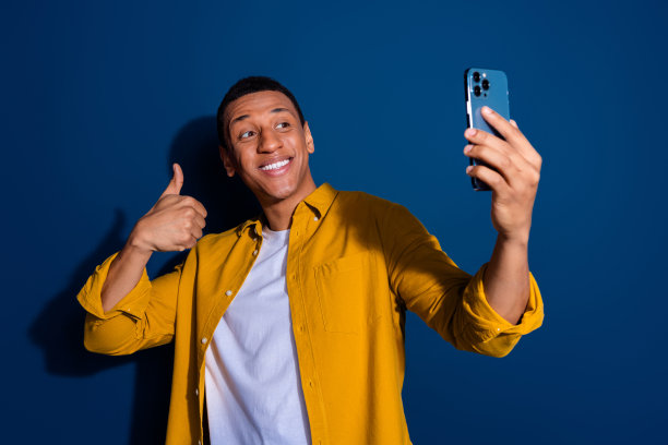 年轻的博主男孩穿着黄色时髦衬衫的照片，正面照片显示拇指向上孤立在深蓝色的背景图片下载