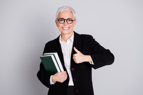 漂亮的高级女专业讲师的照片肖像拿着书拇指向上穿正装眼镜孤立在灰色背景图片下载