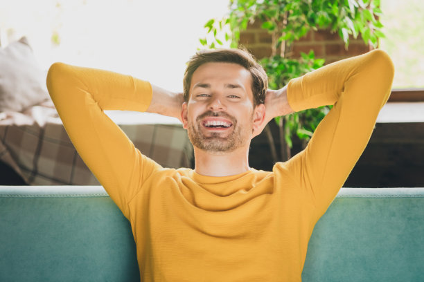 漂亮年轻人的照片肖像放松双手在头后面，坐在沙发上，穿着黄色的衣服现代室内平坦的室内图片下载