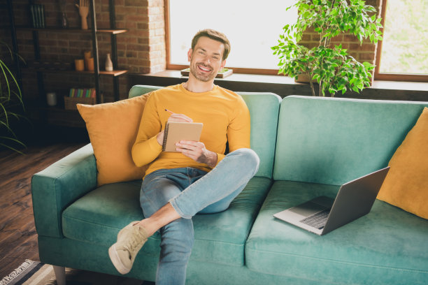 漂亮年轻人的照片肖像坐在沙发上上网学习写笔记本穿着黄色的衣服现代室内平坦的室内图片下载