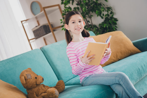 年轻的孩子的照片，黑发女孩呆在家里度过暑假周末，在沙发上的泰迪熊，独自在她的房间里看书图片下载