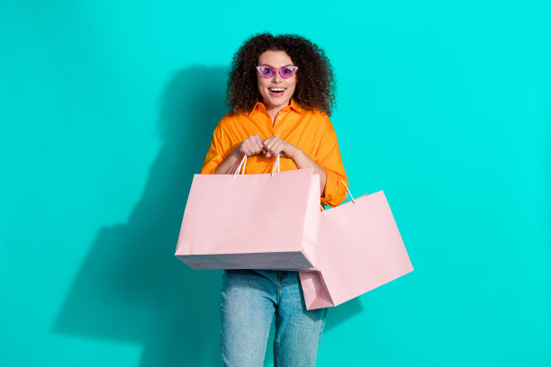 印象深刻的积极的好女人穿着橙色衬衫拿着购物袋惊讶地盯着孤立的蓝绿色背景的照片图片下载
