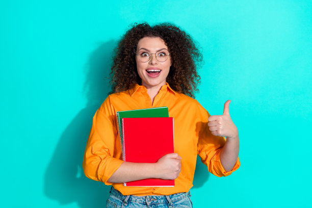 印象深刻的女人的照片，波浪的头发穿着衬衫，戴着眼镜，拿着书，大拇指向上翘，赞成孤立在蓝绿色的背景上图片下载